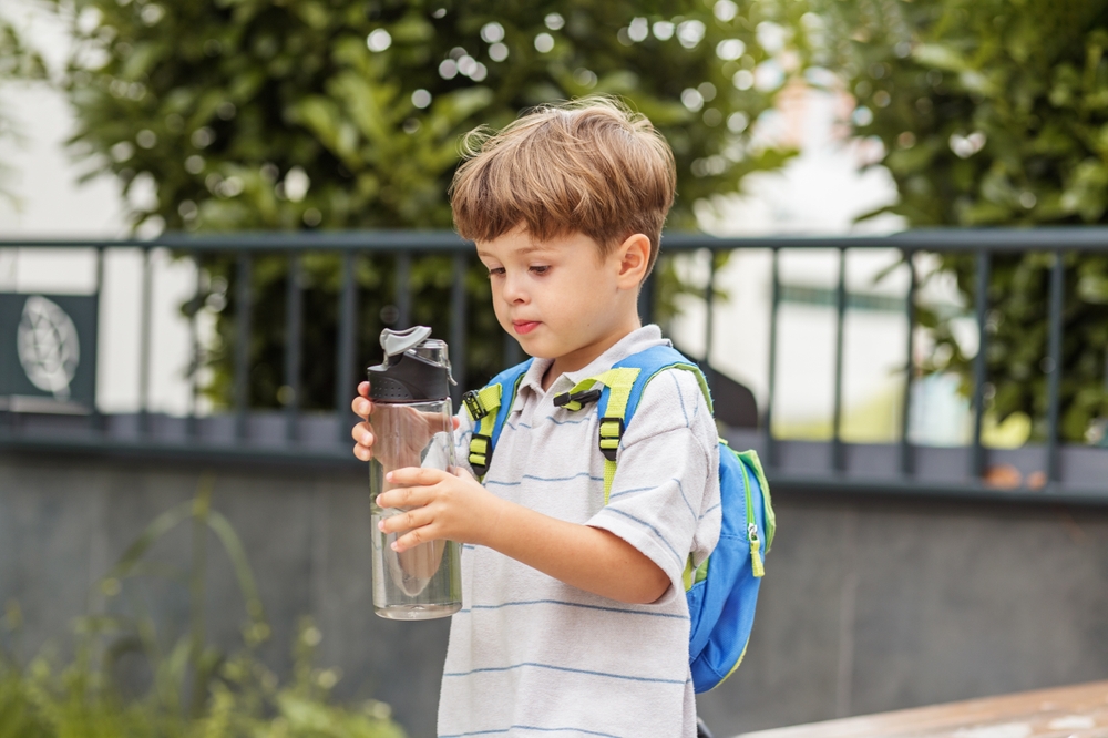 A child with a small backpack on, drinking water from a clear bottle.