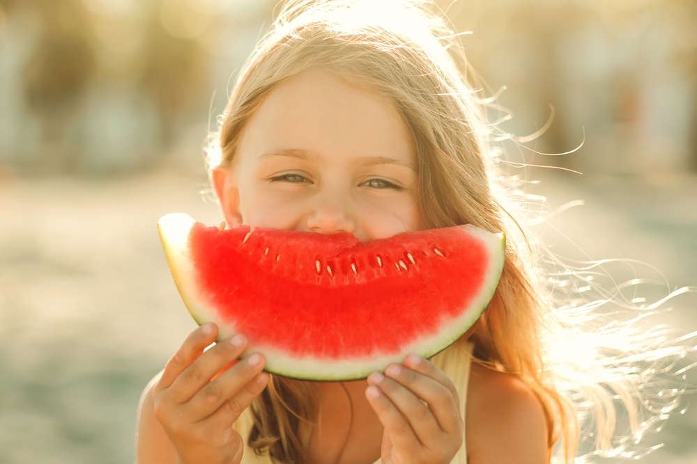 A child holding a slice of watermelon in front of their face as though it is a huge grin, indicating healthy food for oral health creates healthy smiles.