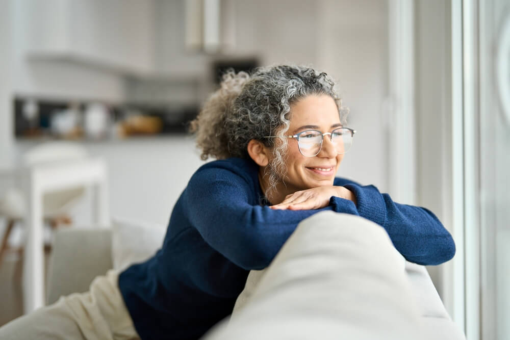 A grey-haired, middle-aged woman smiling as she leans on her couch, glad she saw a dentist in Arana Hills about women's health and its impact on her oral health.