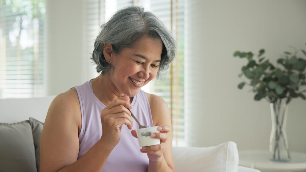 A grey haired middle-aged woman smiling and eating yogurt.