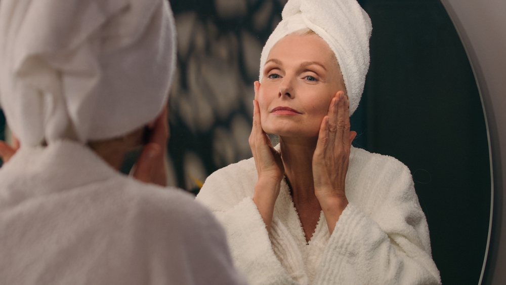 A woman with her hands on her cheeks looking into her bathroom mirror wearing a towel on her head and in a bathrobe.
