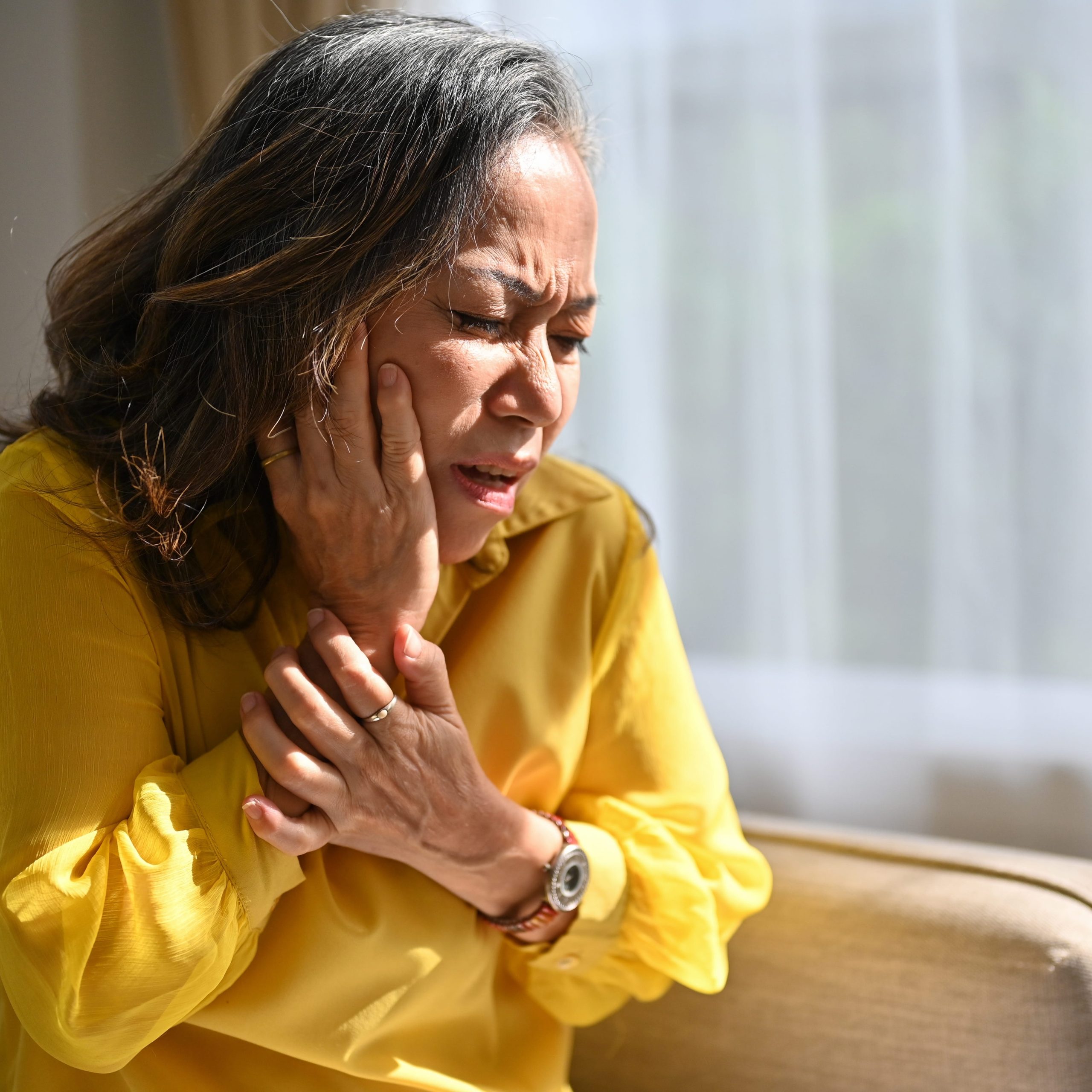 A mature woman sitting indoors holding her jaw in discomfort, highlighting the experience of menopause jaw pain. 