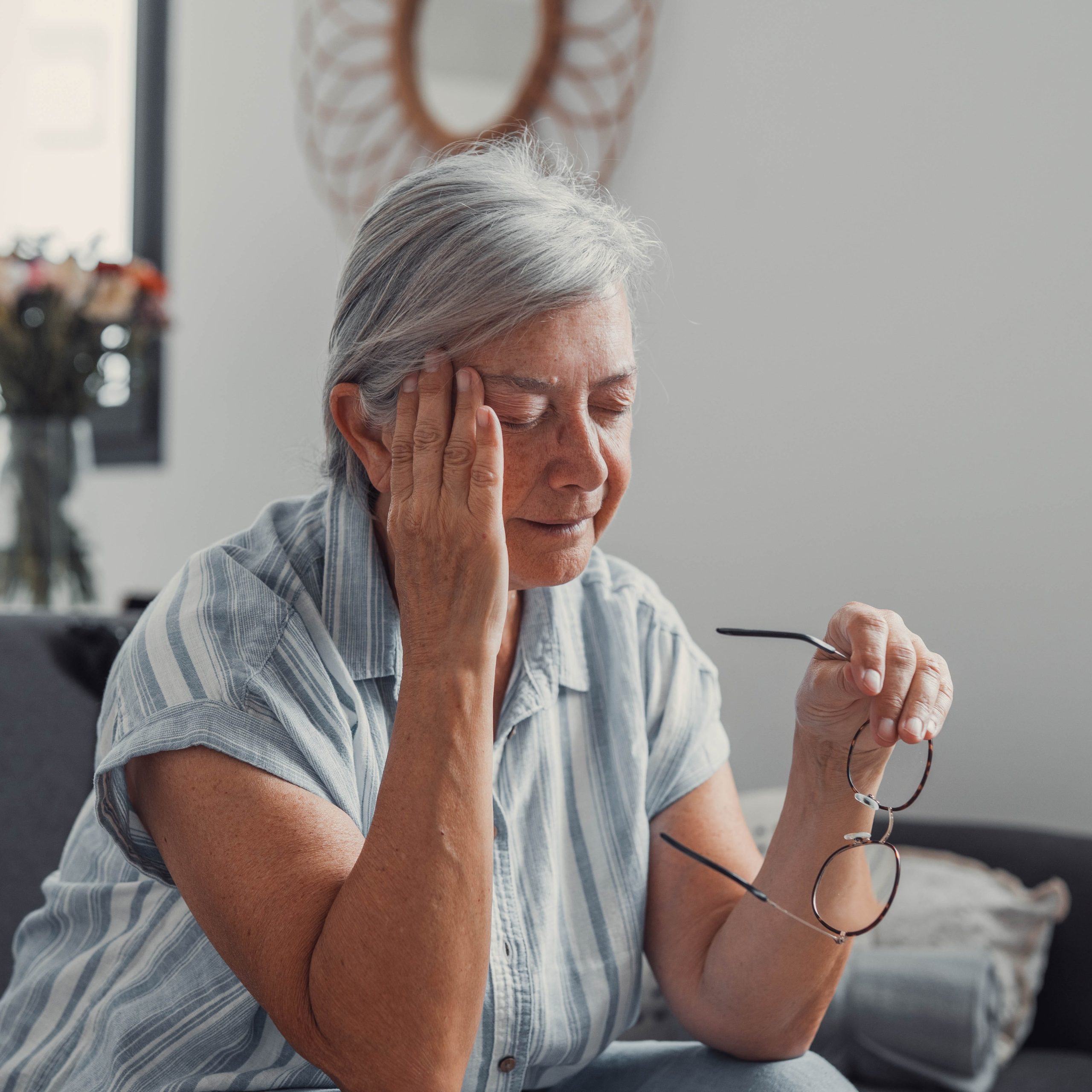 An older woman sitting on a couch holding her glasses and touching her temple, showcasing a headache related to menopause and TMJ.