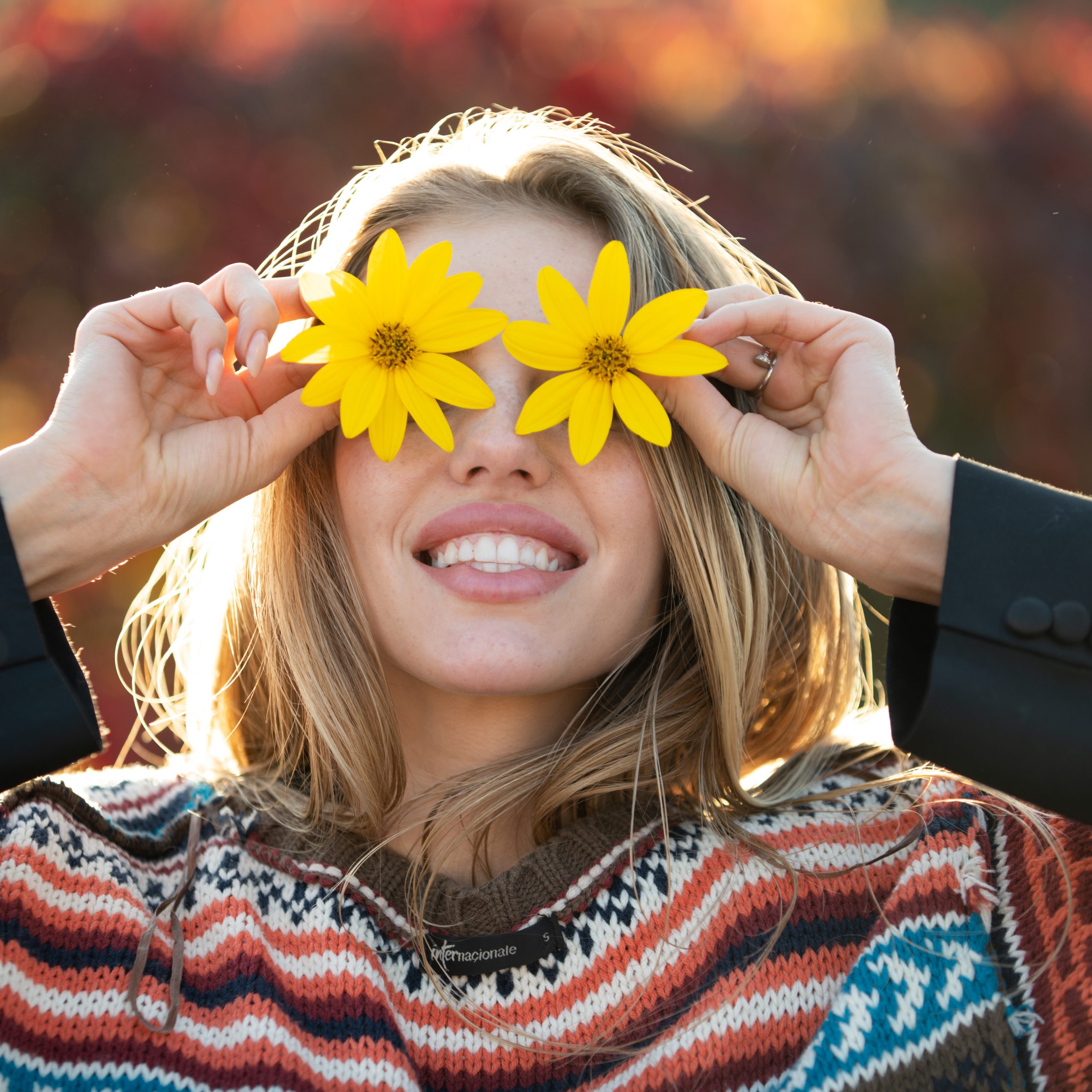 A smiling woman with a radiant, refreshed look holding bright yellow flowers over her eyes during a sunny spring day.